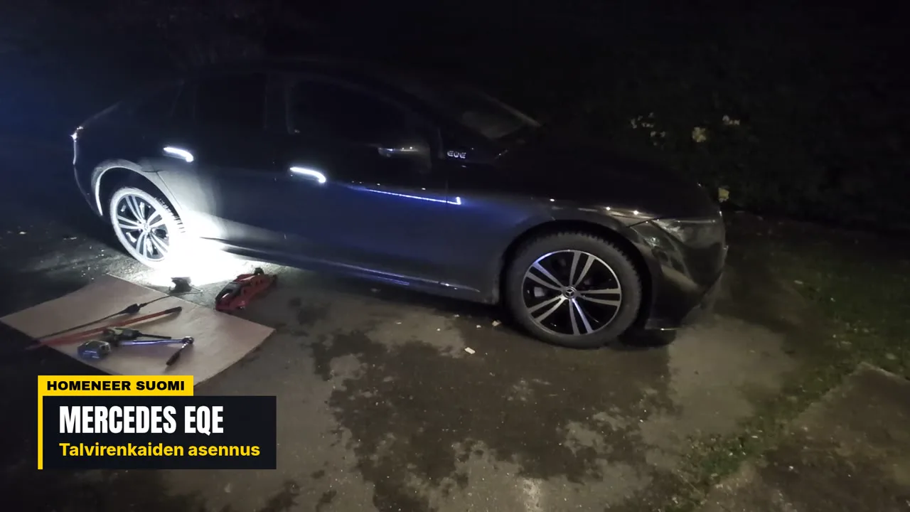 Dark Mercedes-Benz EQE sedan at night during a winter-wheel change, with a floor jack and breaker bar on the ground